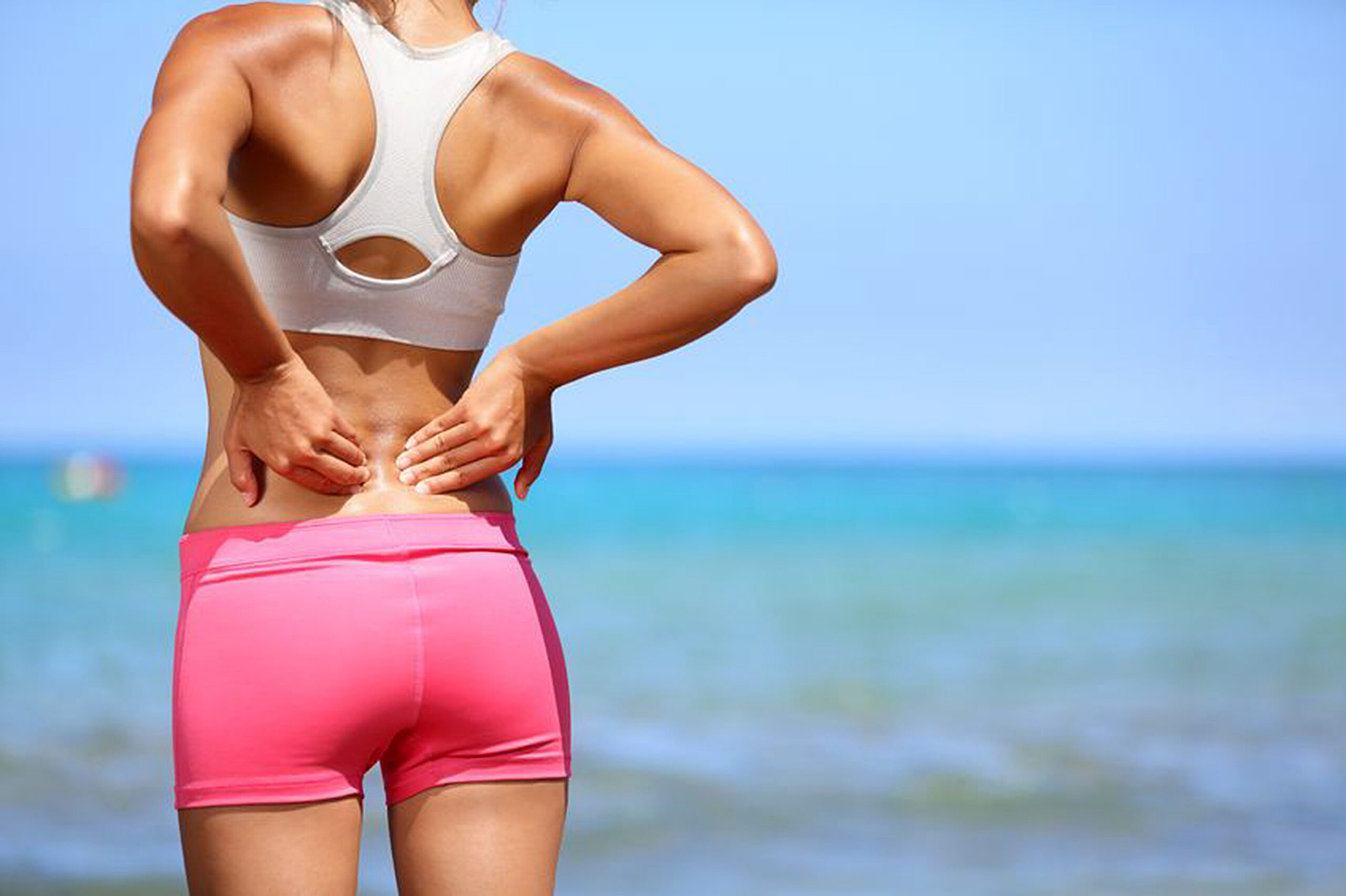 20996111 - back pain. athletic woman in pink sportswear standing at the seaside rubbing the muscles of her lower back, cropped torso portrait.
